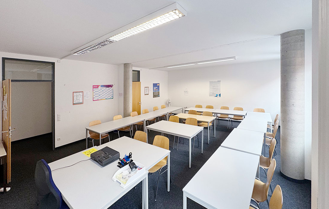 Leerer Unterrichtsraum im IB Sprachinstitut Böblingen, die Tische stehen in einer U-Form mit Tischen dazwischen. Auf dem Lehrertisch liegen Unterrichtsutensilien. Die Tür steht offen.