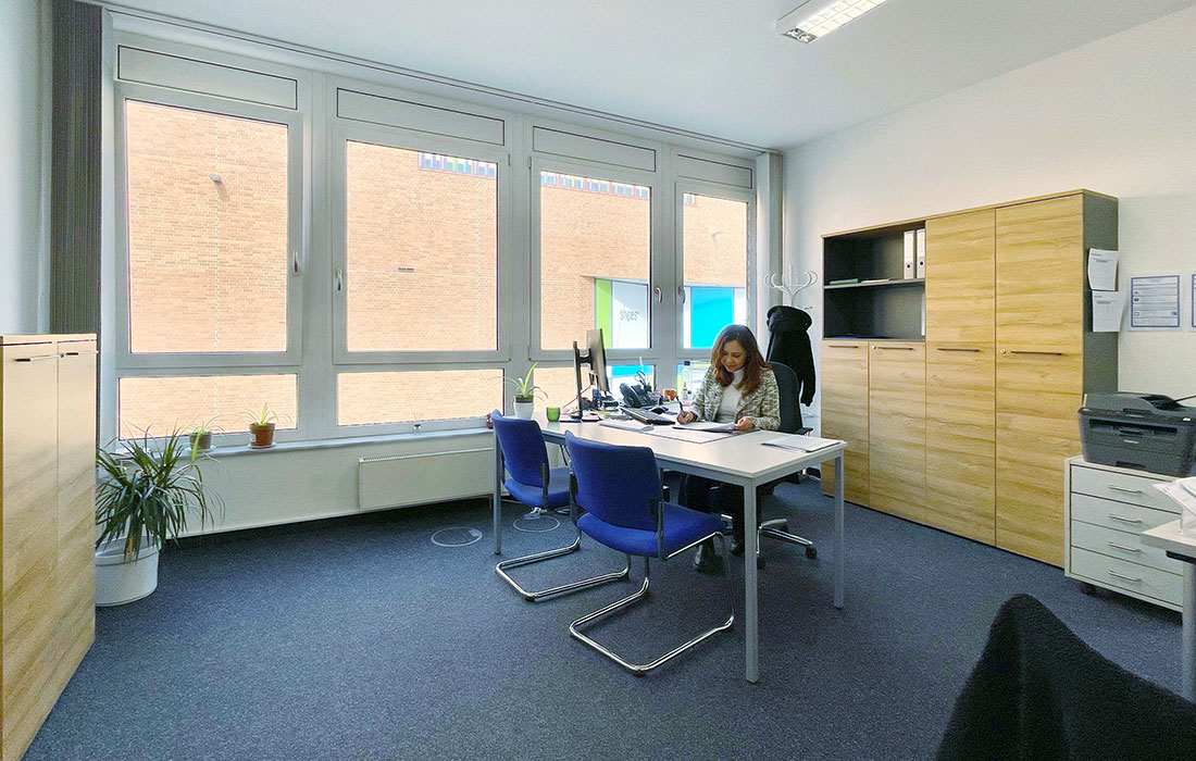 Ein Büroraum mit Büromöbeln aus Holz an der linken und rechten Wand. Dazwischen steht ein Schreibtisch mit zwei Stühlen für Besucher, am Schreibtisch arbeitet eine Frau